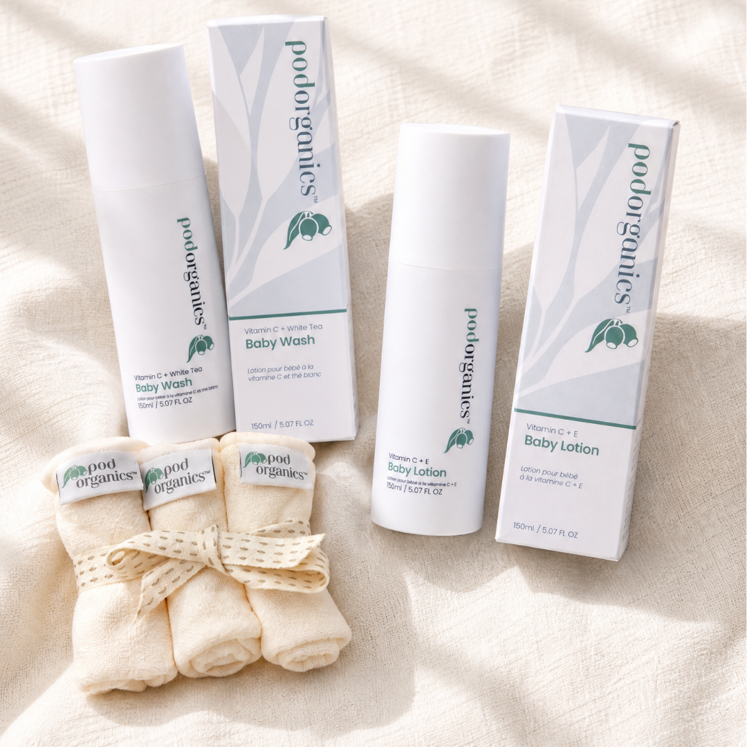 Pod Organics baby care set on a beige fabric background, including a Baby Wash bottle and box, a Baby Lotion bottle and box, and three rolled cream baby towels tied with a ribbon.