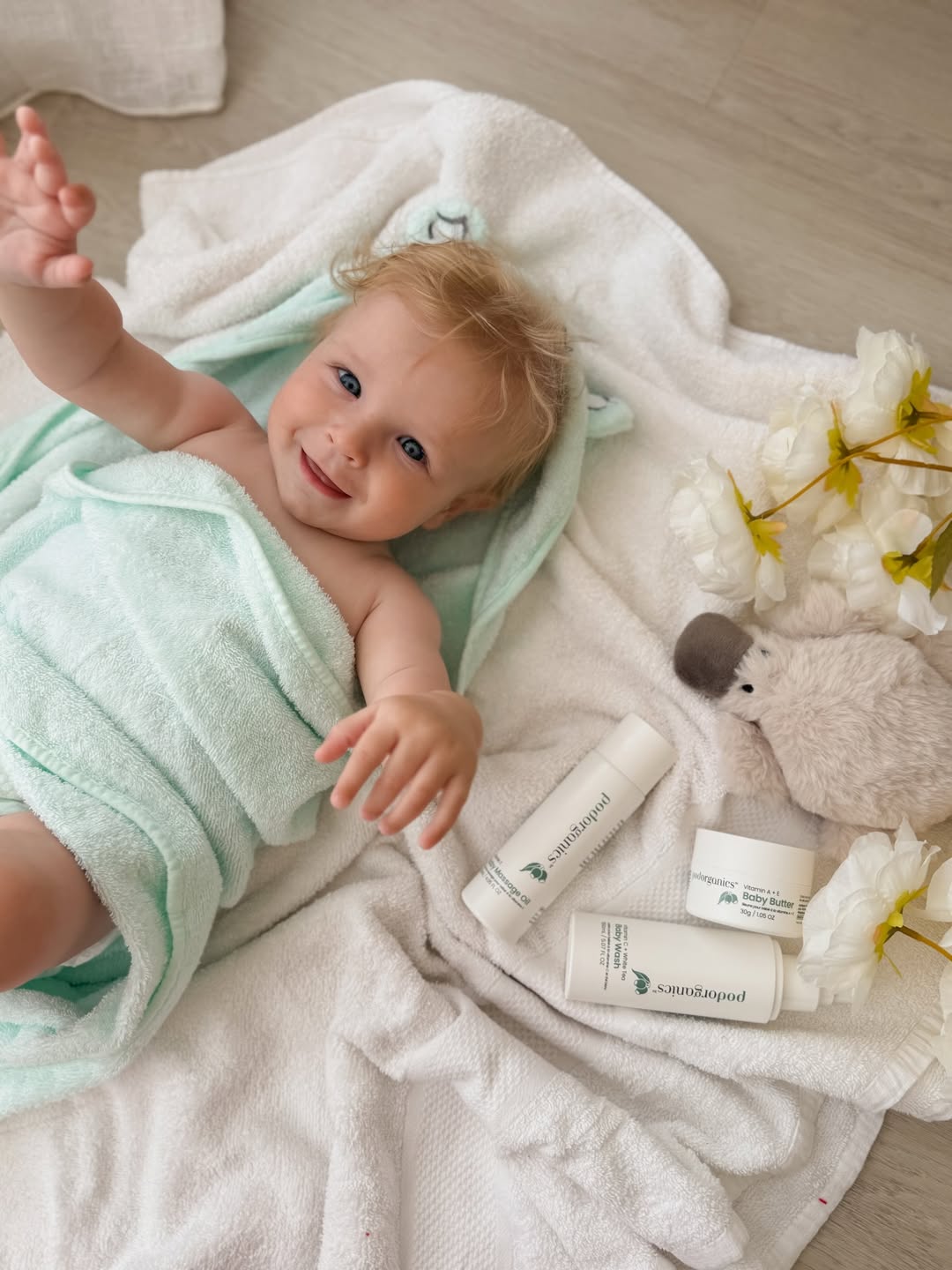 A baby wrapped in a mint green towel lies on a white blanket with three Sandipod skincare tubes and a plush toy nearby, on a light wood floor.