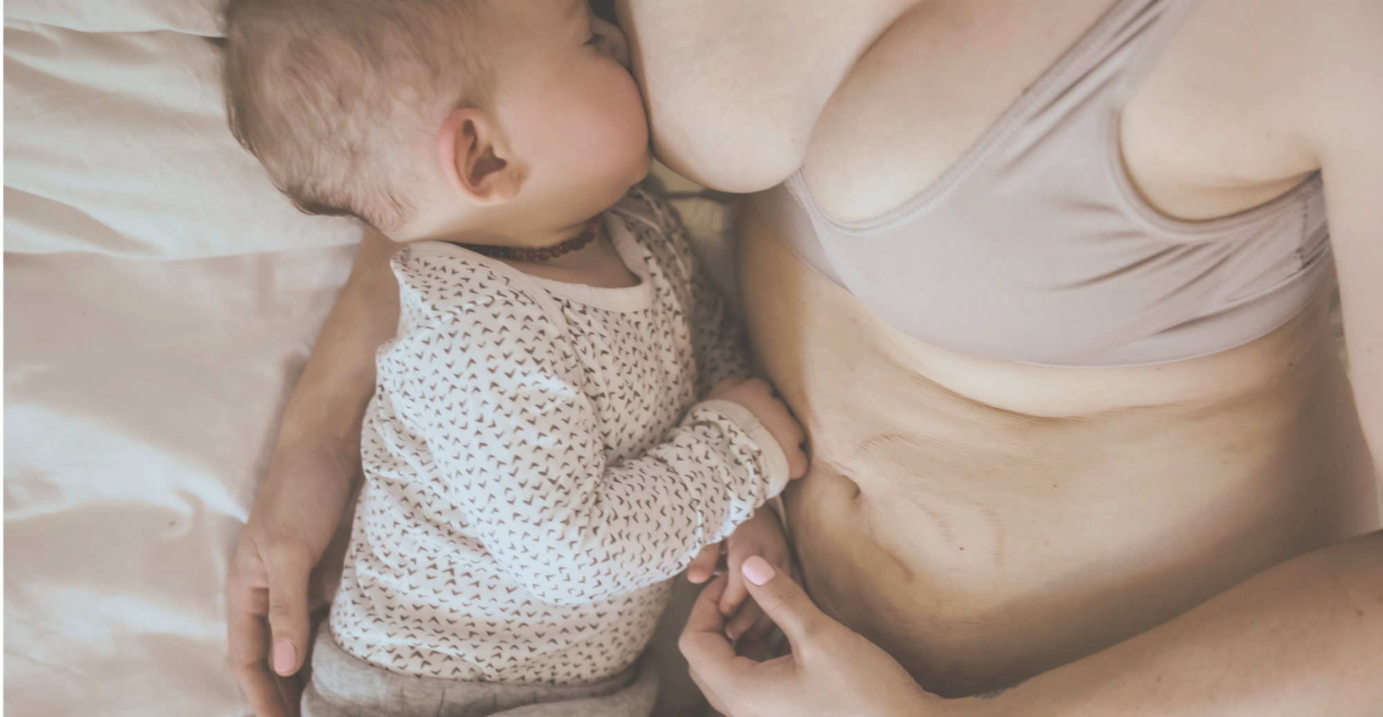 A close-up lifestyle shot of a baby nursing from a mother's breast while lying on a bed. The baby is wearing a cream-colored long-sleeve shirt with small brown polka dots, and the mother is wearing a beige nursing bra.