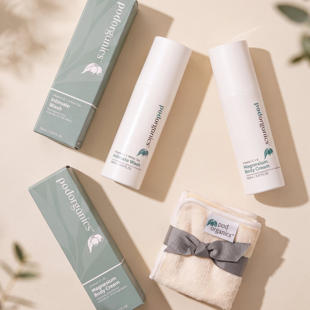 A flat-lay of Pod Organics body care products on a warm beige background with soft leaf shadows. The set includes two white tubes of intimate wash and magnesium body cream, two sage green boxes, and a folded cream towel tied with a gray ribbon.