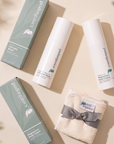 A flat-lay of Pod Organics body care products on a warm beige background with soft leaf shadows. The set includes two white tubes of intimate wash and magnesium body cream, two sage green boxes, and a folded cream towel tied with a gray ribbon.